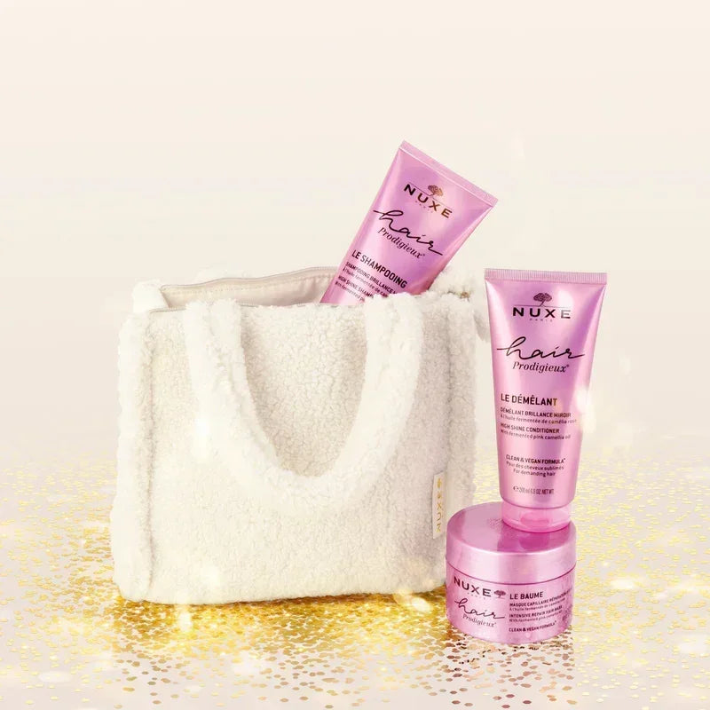 Nuxe luxury hair care set with shampoo, conditioner, and balm in pink packaging, next to a white plush bag on a gold sparkling background, sold on Goldfarmaci Albania.